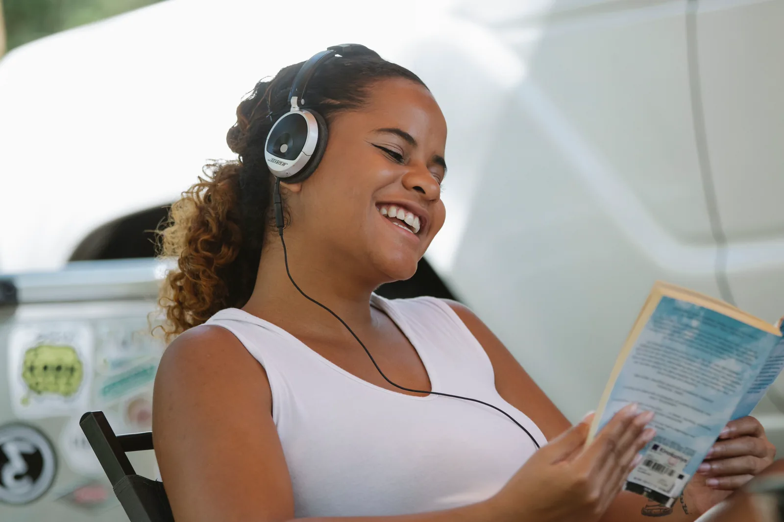 Mujer sonriente escucha con auriculares mientras lee un libro al aire libre.